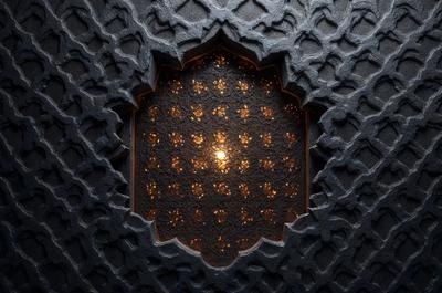 The primary subject of the image is a light shining through a window with a patterned design. The setting is a room with a stone wall, and the visual style appears to be a combination of a photo and a digital art or illustration. The colors and mood of the image are predominantly dark, with the light source creating a contrasting effect.
