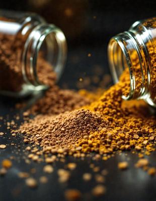 The primary subject of the image is a close-up of a spice, likely turmeric, with a yellow color. The spice is being poured into a glass jar, which is placed on a black surface. The setting appears to be a kitchen or a similar environment where spices are commonly used. The visual style of the image is a close-up, which emphasizes the details of the spice and the jar. The colors and mood of the image are warm and inviting, as the yellow spice and the
