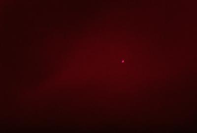 The primary subject of the image is a red background with a red dot in the center. The setting appears to be a dark room, and the visual style is a close-up of the red dot. The colors and mood of the image are predominantly red, creating a bold and striking appearance.