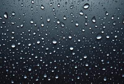 The primary subject of the image is a rainy day, as evidenced by the numerous raindrops falling on a dark background. The setting is a black sky, which contrasts with the raindrops, creating a visually striking scene. The visual style is a close-up of the raindrops, emphasizing their individual shapes and sizes. The colors and mood of the image are predominantly dark, with the raindrops being the main source of light and contrast.