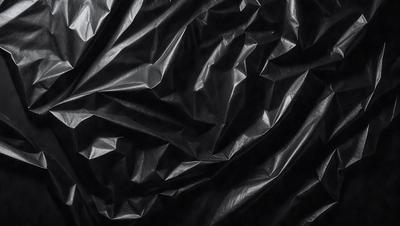 The primary subject of the image is a close-up of a black plastic bag. The setting is a dark background, which emphasizes the plastic bag. The visual style is a black and white photo, giving the image a classic and timeless appearance. The colors and mood of the image are predominantly dark, with the plastic bag being the main focal point.