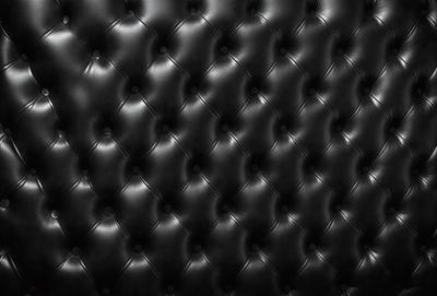 The primary subject of the image is a black leather couch. The setting is a black background, which creates a striking contrast with the couch. The visual style is a close-up of the couch, showcasing its texture and details. The colors and mood of the image are predominantly dark, with the black leather couch being the main focal point.