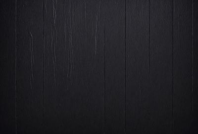 The primary subject of the image is a black wooden wall. The setting is a dark room, and the visual style is a close-up of the wooden wall. The colors and mood of the image are predominantly dark, with the black wooden wall being the main focus.