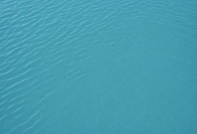 The primary subject of the image is a large body of blue water, which appears to be a lake. The water is calm and serene, with no visible disturbances or ripples. The setting is a clear day, with the sky visible above the water. The visual style of the image is a close-up of the water, showcasing its depth and tranquility. The colors and mood of the image are predominantly blue, creating a sense of calmness and relaxation.