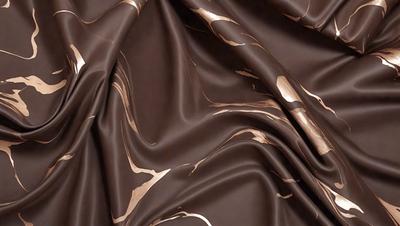 The primary subject of the image is a brown fabric with gold and brown patterns. The setting is a close-up view of the fabric, showcasing its intricate design. The visual style is a detailed, textured image that highlights the fabric's unique pattern. The colors and mood of the image are warm and earthy, with the gold and brown tones creating a sense of luxury and sophistication.
