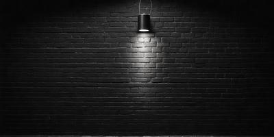 The primary subject of the image is a light fixture mounted on a brick wall. The light is shining brightly, illuminating the dark brick wall. The setting is a black brick wall, which serves as the background for the light fixture. The visual style of the image is a photo, capturing the light and the brick wall in detail. The colors and mood of the image are predominantly dark, with the light fixture being the main source of illumination.