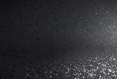 The primary subject of the image is a close-up of a black surface with a pattern of glittering specks. The setting is a dark room, and the visual style appears to be a photo or a digital artwork. The dominant colors are black and the glittering specks, which create a moody and intriguing atmosphere.