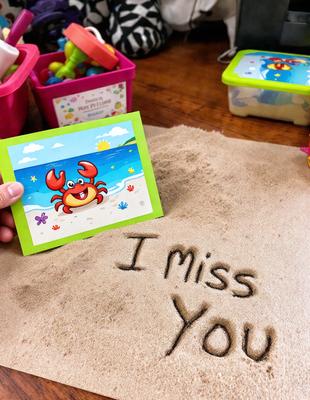 The primary subject of the image is a person holding a card with a crab on it. The card is placed on a sandy surface, which is the setting for the scene. The visual style of the image is a combination of photo and digital art, as it appears to be a hand-drawn illustration. The colors and mood of the image are warm and inviting, with the sandy surface and the crab card creating a sense of relaxation and enjoyment.