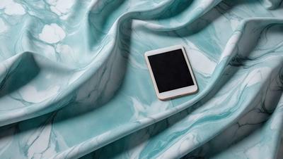 The primary subject of the image is a cell phone placed on a blue and white marbled surface. The setting is a bedroom, with the bedspread and sheets visible in the background. The visual style is a photo, capturing the cell phone and its surroundings. The colors and mood of the image are predominantly blue and white, creating a calm and serene atmosphere.