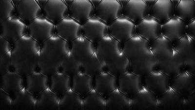 The primary subject of the image is a black leather couch. The setting is a black background, which creates a striking contrast with the couch. The visual style is a close-up of the couch, showcasing its texture and details. The colors and mood of the image are predominantly black, giving it a sleek and modern appearance.