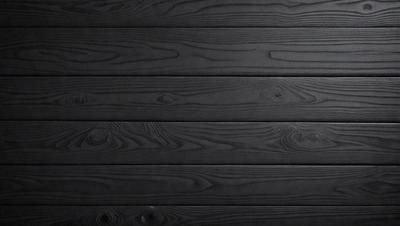 The primary subject of the image is a wooden wall. The setting is a black background, which creates a striking contrast with the wooden surface. The visual style is a close-up of the wooden wall, showcasing its texture and details. The colors and mood of the image are predominantly black, giving it a bold and dramatic appearance.
