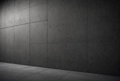 The primary subject of the image is a person standing in front of a large concrete wall. The setting is an outdoor environment, with the person standing on a sidewalk. The visual style of the image is a black and white photo, which adds a sense of drama and contrast to the scene. The dominant colors are black and white, and the overall atmosphere is somewhat moody and dramatic.