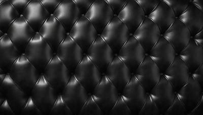 The primary subject of the image is a black leather couch. The setting is a room with a black background, which creates a striking contrast with the couch. The visual style is a close-up of the couch, showcasing its texture and details. The colors and mood of the image are predominantly black, giving it a sleek and modern appearance.