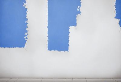 The primary subject of the image is a blue wall with peeling paint. The setting is a room with a white floor. The visual style is a photo of the wall, showcasing the peeling paint and the blue color. The colors and mood of the image are predominantly blue and somewhat melancholic due to the peeling paint.