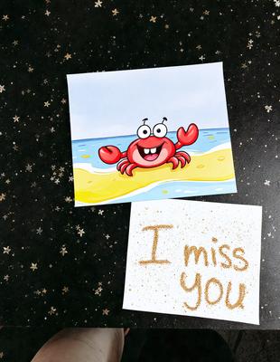 The primary subject of the image is a crab, which is the main focus of the scene. The crab is depicted on a card, and it appears to be smiling. The setting is a beach, with sand visible in the background. The visual style of the image is a combination of a photo and a drawing, creating a unique and playful appearance. The colors and mood of the image are bright and cheerful, with the crab's smile adding a sense of warmth and happiness to the scene.