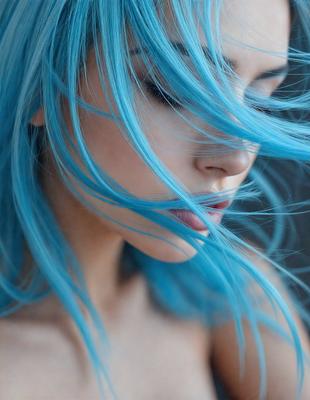 The primary subject of the image is a woman with blue hair. She is looking down and has her hair blowing in the wind. The setting is outdoors, with a background of a sky. The visual style is a close-up of the woman's face, and the colors are predominantly blue, giving the image a calm and serene atmosphere.