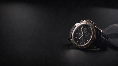 The primary subject of the image is a watch, which is prominently displayed in the foreground. The setting is a dark background, which contrasts with the watch and adds a sense of depth to the image. The visual style is a close-up of the watch, showcasing its details and design. The colors and mood of the image are predominantly dark, with the watch being the main focal point.
