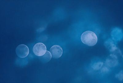 The primary subject of the image is a group of five moons, each with a different size and position, floating in the sky. The setting is a cloudy blue sky, which serves as the backdrop for the moons. The visual style is a combination of photo and digital art, creating a unique and captivating scene. The colors and mood of the image are predominantly blue, with the moons and the sky creating a serene and peaceful atmosphere.