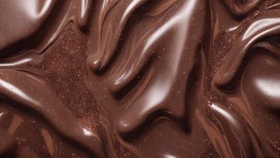 The primary subject of the image is a close-up of a chocolate cake with a swirled pattern. The setting is a table, and the visual style is a close-up photo. The colors and mood of the image are predominantly brown, with a rich and indulgent atmosphere.
