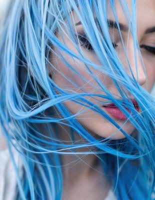 The primary subject of the image is a woman with blue hair, who is looking upward and has her eyes closed. She is wearing lipstick and has a blue-green hair color. The setting is outdoors, with the woman standing in front of a blue sky. The visual style is a close-up of the woman's face, and the colors and mood are predominantly blue and serene.