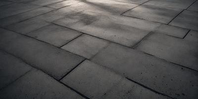 The primary subject of the image is a large, square, grey brick tile floor. The setting is an outdoor area with a sunny day, as the sunlight shines on the floor. The visual style is a photo, capturing the details of the brick tile floor. The colors and mood of the image are predominantly grey, giving it a calm and serene atmosphere.