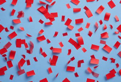 The primary subject of the image is a large number of red paper hearts scattered across a blue background. The hearts are of various sizes and are spread out across the entire image. The setting appears to be a creative and playful environment, possibly for a celebration or artistic project. The visual style of the image is a combination of photo and digital art, creating a vibrant and lively atmosphere. The dominant colors are red and blue, with the red paper hearts standing out against the blue background.