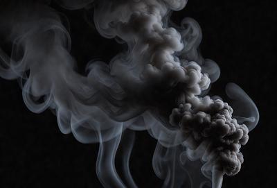 The primary subject of the image is a large, dark, and smoky cloud. The setting is a black background, which emphasizes the cloud's shape and form. The visual style is a black and white photo, giving the image a dramatic and moody atmosphere. The colors and mood of the image are predominantly dark and mysterious, with the cloud serving as the focal point of the scene.