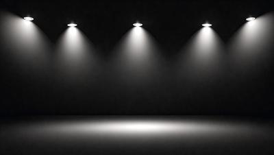 The primary subject of the image is a black background with three bright lights shining on it. The lights are positioned at the top left, top right, and bottom left of the background. The setting is a dark room, and the visual style is a black and white photo. The overall atmosphere is dramatic and moody, with the lights creating a striking contrast against the darkness.