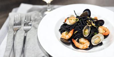 The primary subject of the image is a plate of food, which includes shrimp, scallops, and noodles. The plate is placed on a dining table, and there is a wine glass next to it. The visual style of the image is a photo, and the colors and mood are predominantly black and white, giving the scene a classic and elegant appearance.