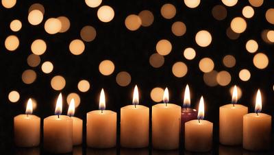 The primary subject of the image is a row of lit candles. The setting is a dark room with a patterned background, creating a visually striking contrast. The visual style is a photo, capturing the glowing candles and their reflections on the patterned background. The colors and mood of the image are predominantly dark, with the candles providing a warm and inviting atmosphere.