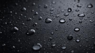 The primary subject of the image is a close-up of a black surface with water droplets on it. The setting is a dark background, and the visual style appears to be a photo. The colors in the image are predominantly black and white, with the water droplets adding a touch of gray. The overall atmosphere is moist and somewhat mysterious.