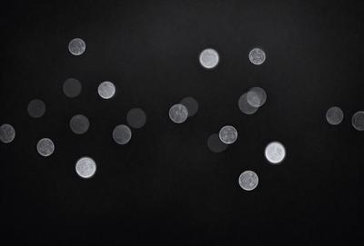 The primary subject of the image is a group of small, round, glowing lights. These lights are scattered across a dark background, creating a visually striking scene. The visual style of the image is a combination of a photo and digital art, as the lights appear to be floating in the darkness. The colors and mood of the image are predominantly dark, with the glowing lights providing a sense of warmth and energy.