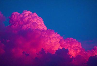 The primary subject of the image is a large, pink cloud with a purple hue. The cloud is floating in a blue sky, creating a striking contrast between the cloud and the sky. The visual style of the image is a photo, capturing the beauty of the cloud in its natural environment. The colors and mood of the image are vibrant and lively, with the pink and purple tones evoking a sense of warmth and tranquility.