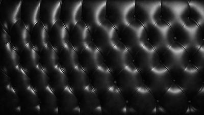The primary subject of the image is a black leather couch. The setting is a room with a black background, which creates a contrast with the couch. The visual style is a close-up of the couch, showcasing its texture and design. The colors and mood of the image are predominantly black, giving it a sleek and modern appearance.