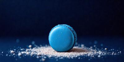 The primary subject of the image is a blue macaroon with white sprinkles on top. The setting is a blue background, which creates a contrast with the blue and white colors of the macaroon. The visual style is a close-up of the macaroon, showcasing its details. The colors and mood of the image are predominantly blue and white, giving it a clean and fresh appearance.