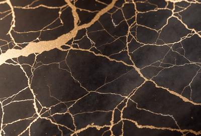 The primary subject of the image is a close-up of a black and gold patterned surface. The setting appears to be a dark room, with the patterned surface being the main focus. The visual style of the image is a combination of a photo and digital art, creating a unique and intriguing appearance. The dominant colors in the image are black and gold, giving the scene a sophisticated and elegant atmosphere.