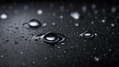 The primary subject of the image is a close-up of water droplets on a black surface. The setting is a dark background, which contrasts with the bright droplets. The visual style is a black and white photo, giving the image a classic and timeless appearance. The colors and mood of the image are predominantly dark, with the water droplets being the main source of light and interest.
