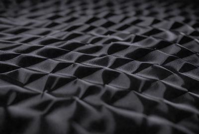 The primary subject of the image is a black fabric with a pattern of small squares. The setting is a dark background, which contrasts with the fabric. The visual style is a close-up of the fabric, showcasing its texture and pattern. The colors are predominantly black, giving the image a moody and dramatic atmosphere.