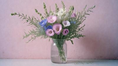 The primary subject of the image is a vase filled with a variety of flowers. The vase is placed on a table, and the flowers are arranged in a visually appealing manner. The setting is indoors, with a pink background visible in the image. The visual style is a close-up of the vase and flowers, showcasing the details of the arrangement. The colors and mood of the image are predominantly warm and inviting, with the flowers adding a touch of natural beauty to the scene.