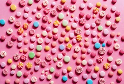 The primary subject of the image is a large pink surface covered with a variety of colorful candy-shaped candies. The candies are arranged in a visually appealing pattern, creating a vibrant and playful atmosphere. The setting is a pink background, which complements the colorful candies. The visual style of the image is a combination of photo and digital art, as it captures the intricate details of the candies while maintaining a visually pleasing composition. The colors and mood of