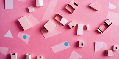 The primary subject of the image is a collection of small, white boxes arranged on a pink background. The setting appears to be a creative workspace or a display of artistic pieces. The visual style of the image is a combination of digital art and illustration, with the boxes being digitally created and placed on the pink surface. The colors and mood of the image are predominantly pink, which gives it a warm and inviting atmosphere.