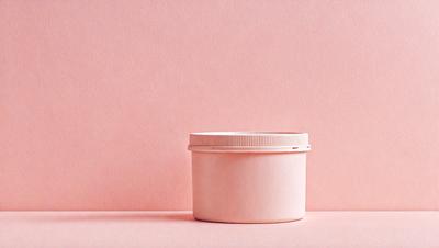 The primary subject of the image is a small, white, plastic container. The container is placed on a pink background, which gives the scene a warm and inviting atmosphere. The visual style of the image is a close-up of the container, showcasing its details and texture. The colors and mood of the image are predominantly pink, creating a sense of softness and femininity.