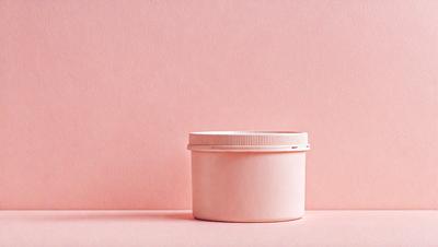The primary subject of the image is a small, white, plastic container with a lid. The container is placed on a pink surface, which serves as the background. The visual style of the image is a photo, capturing the details of the container and its surroundings. The colors in the image are predominantly pink, creating a warm and inviting atmosphere.
