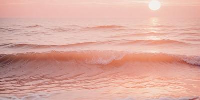 The image features a beautiful sunset over the ocean, with the sun peeking through the clouds. The sky is a mix of pink and orange hues, creating a serene and picturesque scene. The ocean is filled with waves, some of which are crashing onto the shore. The primary subject of the image is the ocean and the sunset, with the setting being the ocean and the background being the sky. The visual style of the image is a natural, realistic representation of the sunset and the ocean. The colors