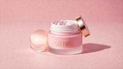 The primary subject of the image is a jar of pink cream, which is placed on a pink surface. The jar is filled with a pink substance, and it is accompanied by a small round object, possibly a ball or a marble, which is located next to the jar. The setting appears to be a simple and elegant display of the pink cream, with the pink surface providing a cohesive and visually appealing backdrop. The visual style of the image is a close-up of the jar