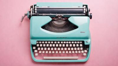 The primary subject of the image is a blue typewriter with a white keyboard. The setting is a pink surface, which provides a contrasting background for the typewriter. The visual style is a close-up of the typewriter, showcasing its details and design. The colors and mood of the image are predominantly blue and pink, creating a nostalgic and vintage atmosphere.