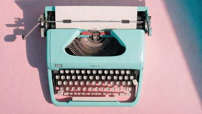 The primary subject of the image is a blue typewriter with a white keyboard. The setting is a pink background, which adds a contrasting color to the scene. The visual style is a close-up of the typewriter, showcasing its details. The colors and mood of the image are predominantly pink and blue, creating a calm and nostalgic atmosphere.
