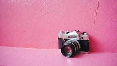 The primary subject of the image is a silver camera, which is placed on a pink wall. The setting is a simple, clean environment with a pink wall as the background. The visual style is a realistic representation of the camera and its surroundings. The colors and mood of the image are predominantly pink, creating a warm and inviting atmosphere.