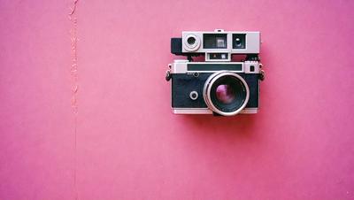 Primary subject: A vintage camera with a black and white lens.

Setting/background: A pink background.

Visual style: The image is a photo of the camera.

Colors and mood: The camera has a black and white lens, and the pink background creates a nostalgic and vintage atmosphere.