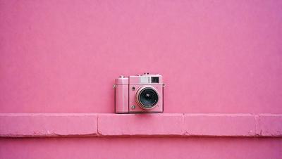The primary subject of the image is a pink camera, which is placed on a pink wall. The setting is a simple, monochromatic environment, with the pink wall serving as the background. The visual style is a photograph, capturing the camera's details and the pink color of the wall. The colors and mood of the image are predominantly pink, creating a warm and inviting atmosphere.